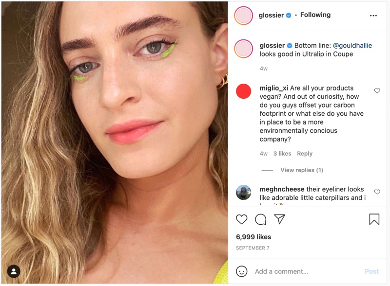 Woman with wavy hair wearing Glossier makeup, featuring vibrant green eyeliner, smiling directly at the camera, highlighting customer engagement in product feedback and community involvement.