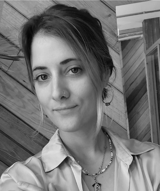 Mujer sonriente con cabello claro y aretes grandes, vestida con camisa clara, en un entorno de madera, representando creatividad y profesionalismo en marketing digital.